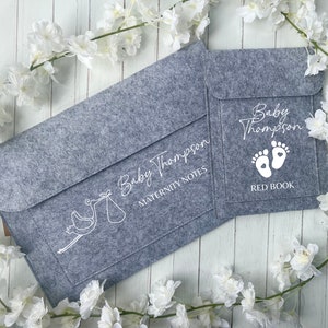 May include: Grey felt folder with a white stork design and the text "Baby Thompson Maternity Notes". A smaller grey felt folder with a white footprint design and the text "Baby Thompson Red Book" is attached to the larger folder.