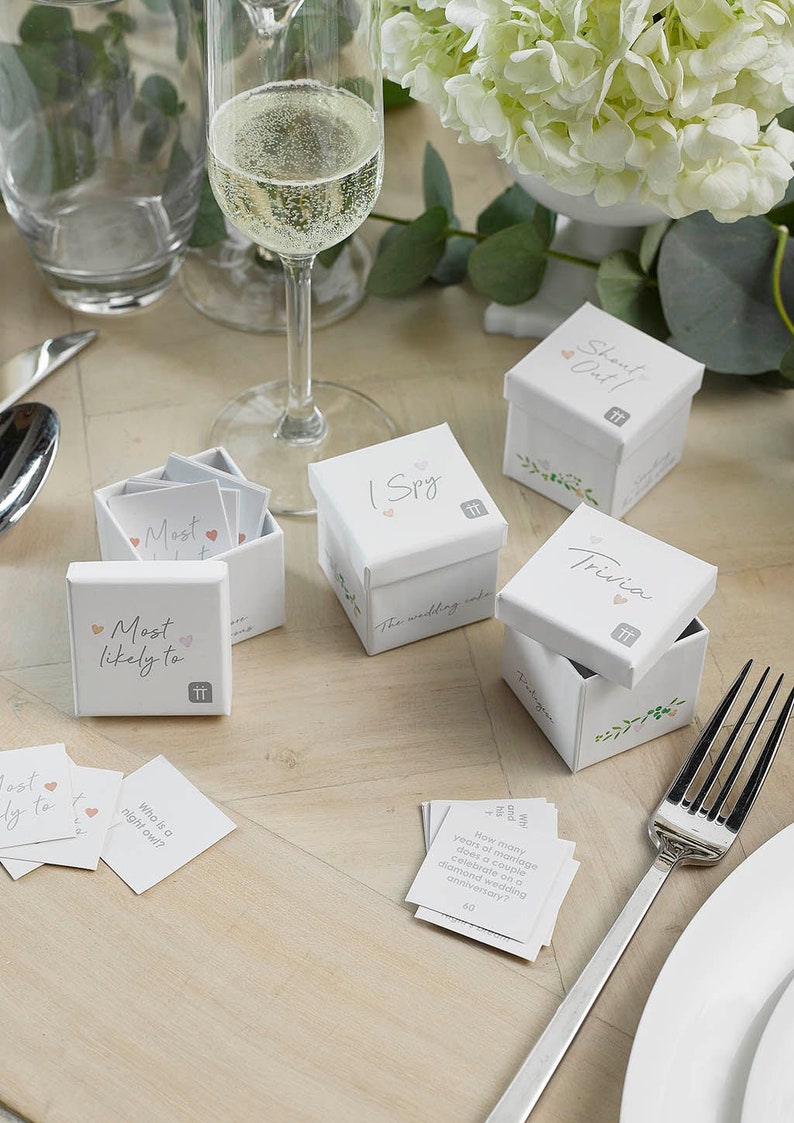 May include: White boxes with floral designs and the text "Most Likely To", "I Spy", "Trivia", and "Shant Out!" on them. The boxes contain cards with questions for a wedding game.