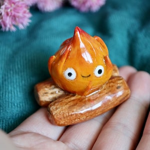 May include: A small, orange, clay figurine of a fire with a smiling face. The fire is sitting on two brown, log-shaped pieces of clay.