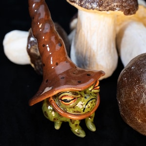 Könnte beinhalten: Eine Keramik-Froschfigur, die einen braunen Hut mit weißen Punkten trägt. Der Frosch hat einen grünen Körper mit braunen Flecken und orangefarbene Augen. Der Frosch sitzt auf einer schwarzen Oberfläche mit zwei braunen Pilzen dahinter.
