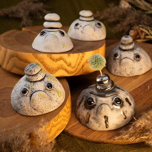 May include: Five ceramic figurines with whimsical faces and stacked stones on their heads. The figurines are painted in shades of gray and white with green accents. One figurine has a small green plant growing from its head.