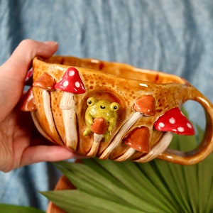 Könnte beinhalten: Eine Keramiktasse in Form von Pilzen mit einem Frosch, der einen Pilz an der Seite hält. Die Tasse ist braun mit roten und weißen Akzenten.