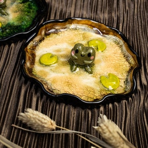 Puede incluir: Dos pequeños platos de cerámica con un borde ondulado, decorados con un efecto de glaseado craquelado. Un plato presenta una figurita de rana verde sobre un nenúfar amarillo, mientras que el otro presenta una figurita de rana verde sobre un nenúfar verde. Ambos platos están sobre una superficie texturizada marrón.