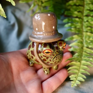 Puede incluir: Figura de rana de cerámica con sombrero de copa marrón. La rana es verde con manchas marrones y ojos naranjas. El sombrero tiene lunares blancos. La figura se sostiene en una mano.