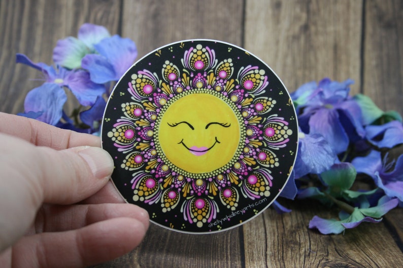 May include: A round sticker with a black background and a yellow smiling sun with pink eyelashes and lips. The sun is surrounded by a pink and gold mandala design.