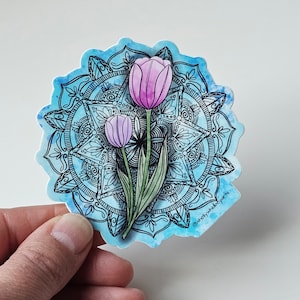 May include: A sticker featuring a mandala design with two pink tulips in the center. The mandala is black and white, and the background is a blue watercolor wash. The sticker has the text "@shellywhidbeyart" in the bottom right corner.