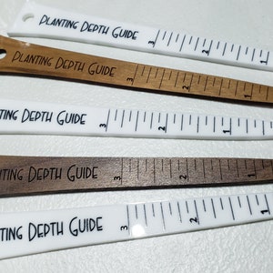 May include: Five planting depth guides, two white and three brown, with a ruler marked in inches from 1 to 3. Each guide has a hole at the top and the text "PLANTING DEPTH GUIDE" is engraved on the front.
