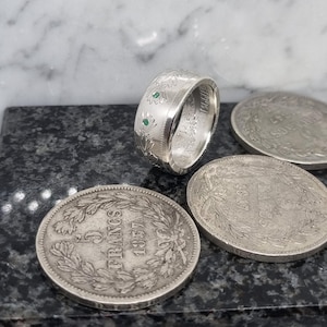 Puede incluir: Anillo de plata hecho con una moneda de 5 Francos con dos piedras preciosas verdes engastadas en la banda.