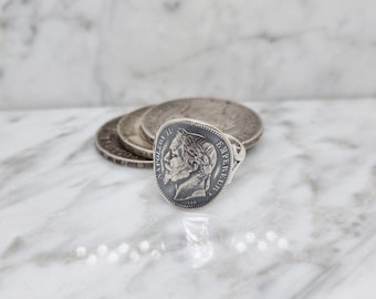 May include: Silver ring featuring a vintage coin with a portrait of a man. The coin is set in a silver band and has a raised edge.