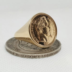 Könnte beinhalten: Ein goldener Ring mit einem Münzendesign, das ein Profil eines Mannes zeigt. Der Ring liegt auf einer Silbermünze.