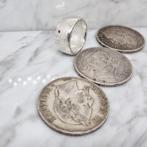 Peut inclure: Une bague en argent avec une pierre rouge sertie dans la bande. La bague est faite d'une pièce d'argent avec l'inscription "NAPOLEON III" visible. La bague est présentée sur une surface en marbre avec trois pièces d'argent.