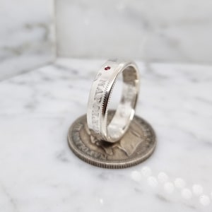 Peut inclure: Bague en argent avec une pierre rouge sertie dans la bande. La bague est gravée des mots "IN GOD WE TRUST".