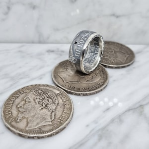 May include: Silver ring made from a vintage French coin featuring the inscription "Napoleon III." The ring is set with a black stone and is displayed on top of three other vintage coins.