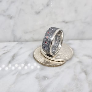 Peut inclure: Une bague en argent fabriquée à partir d'une pièce de monnaie avec une finition oxydée noire. La bague est décorée de pierres précieuses rouges et bleues et d'un motif floral.