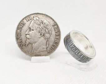 May include: A silver coin featuring a profile of Napoleon III next to a silver ring. The ring is engraved with the word "EMPEREUR". Both items are displayed on clear stands against a white background.