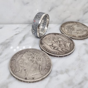 Peut inclure: Une bague en argent avec texte gravé et une petite pierre rouge, accompagnée de plusieurs pièces de monnaie anciennes en argent. La bague porte l'inscription "NAPOLEON III EMPEREUR FRANCAIS". Les pièces présentent des portraits et des inscriptions.
