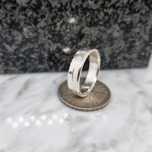 Puede incluir: Un anillo de plata con una banda texturizada y una pequeña piedra roja engastada en la banda. El anillo se muestra junto a una moneda de 20 céntimos de euro para comparar el tamaño.