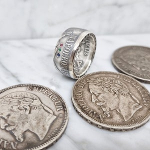 Peut inclure: Une bague en argent fabriquée à partir d'une pièce de monnaie de Napoléon III avec trois pierres précieuses serties dans la bande. La bague est présentée à côté de trois autres pièces de monnaie de Napoléon III.