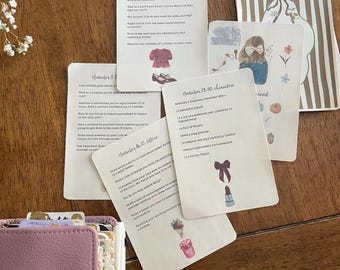 May include: A collection of rectangular journaling prompt cards with printed text and watercolor illustrations, including a pink cardigan, a lipstick, and a person with a bow. A mauve leather-bound planner with white lace trim sits nearby.