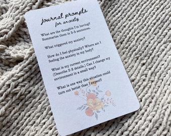 May include: A white card with black text and a floral illustration. The card features journal prompts for anxiety, including questions about thoughts, triggers, physical sensations, and environment.
