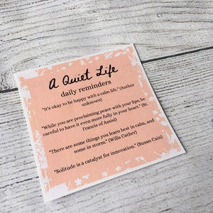 Pu&ograve; includere: Un biglietto rosa e bianco con il testo "A Quiet Life daily reminders" e diverse citazioni sulla pace, la solitudine e l'apprendimento.