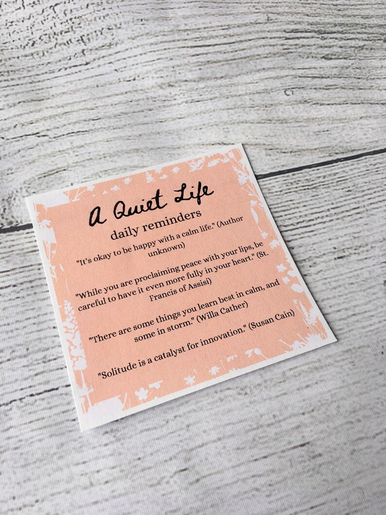 Pu&ograve; includere: Un biglietto rosa e bianco con il testo "A Quiet Life daily reminders" e diverse citazioni sulla pace, la solitudine e l'apprendimento.