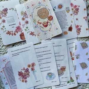 Op de afbeelding: Een verzameling geïllustreerde kaarten met bloemen- en voedselthema's. De kaarten tonen de maand november, een kalender voor 2025 en planningsprompts. Het kleurenpalet omvat roze, beige en groen.