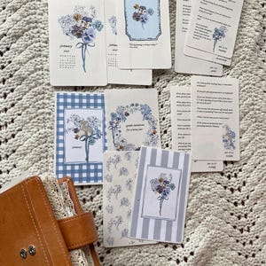 Peut inclure: Un ensemble de cartes à thème floral et un journal en cuir marron. Les cartes présentent des illustrations à l'aquarelle de fleurs, de calendriers et de citations inspirantes. Certaines cartes ont des motifs à carreaux bleus ou à rayures. Le journal est ouvert, révélant un intérieur bordé de dentelle.