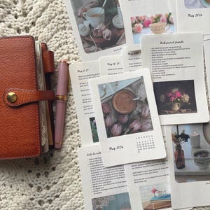 May include: An open brown leather journal with a gold clasp, a pink fountain pen, and several cards with text and images. The cards feature floral and tea-themed illustrations, and journal prompts.