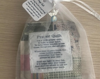May include: A small prayer quilt in a sheer bag, tied with a white ribbon. The quilt has a variety of fabric patches and a tag with text. An angel charm hangs from the ribbon. The text on the tag reads "Prayer Quilt".