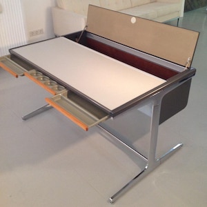 Op de afbeelding: Een mid-century modern bureau met een wit tafelblad en een donkerbruine rand. Het bureau heeft een opklapbaar blad, lades en chromen poten. De open bovenkant onthult een opbergvak. Het bureau is ongeveer 122 cm lang.