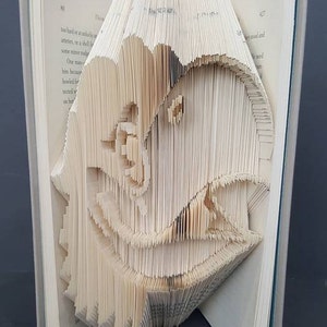 May include: A book with pages folded into a 3D sculpture of a dinosaur head. The sculpture is made from the pages of a book, and the pages are folded to create the shape of the dinosaur's head.