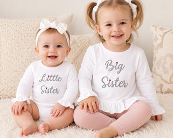 May include: A white long-sleeved bodysuit with the text Little Sister in silver glitter and a small red heart, paired with a white headband. A white long-sleeved ruffled top with the text Big Sister in silver glitter and a small red heart.