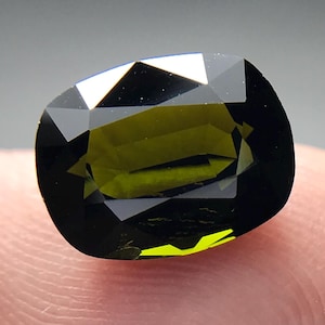 May include: A faceted, cushion-cut gemstone with a dark olive-green hue. The gem is held between fingers, showcasing its polished surfaces and internal reflections. The stone's facets catch the light, creating a play of color.
