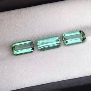 Puede incluir: Tres piedras preciosas verdes, talladas en forma de esmeralda rectangular, se muestran en una caja de terciopelo blanco. Las piedras preciosas están dispuestas en fila, con la piedra más grande a la derecha y la más pequeña a la izquierda.