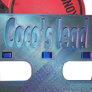 May include: A metallic blue dog lead attachment with the text "Coco's Lead" in a cut-out design. The attachment has a rectangular shape with rounded corners and blue accents. A red and black Kong brand lead is visible.