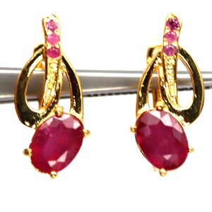 May include: A pair of gold earrings with a ruby gemstone and pink accents. The earrings have a curved design with a small loop at the top.