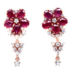 May include: A pair of dangle earrings with a floral design. The earrings feature red ruby stones and clear cubic zirconia stones set in a rose gold tone metal.