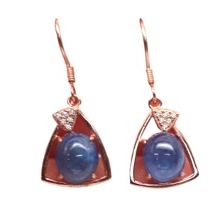 Op de afbeelding: Een paar roségouden oorbellen. Elke oorbel heeft een blauwe edelsteen in een driehoekig frame, geaccentueerd met kleine heldere stenen. De oorbellen hebben een haaksluiting.