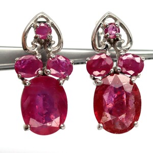 May include: A pair of silver earrings featuring red ruby gemstones. The earrings have a heart-shaped design with three oval-shaped rubies dangling from each heart.