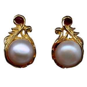 May include: A pair of gold earrings with a pearl and a red gemstone in each earring. The earrings have a delicate, floral design.
