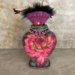 May include: A decorative glass bottle with a floral design and a pink and black feather and doll leg topper. The bottle is decorated with rhinestones.