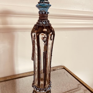 May include: An ornate, clear glass perfume bottle with a blue and bronze color scheme. The bottle features a faceted blue stopper, intricate bronze filigree, and dangling chain accents. The base and neck are adorned with blue gemstones.