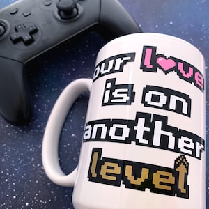 May include: White ceramic mug with a black and white pixelated design that says "Our love is on another level" with a pink pixelated heart and a gold pixelated joystick.