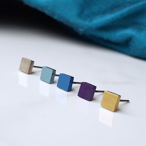May include: Five square-shaped stud earrings in different colors. The earrings are arranged in a row from left to right: gold, light blue, blue, purple, and gold.