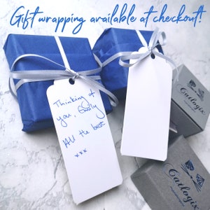 Op de afbeelding: Twee cadeaudozen verpakt in blauw papier met zilveren lint en een wit cadeaukaartje met een handgeschreven bericht. Het cadeaukaartje zegt "Thinking of you, Emily All the best xxx". De dozen staan op een wit oppervlak.