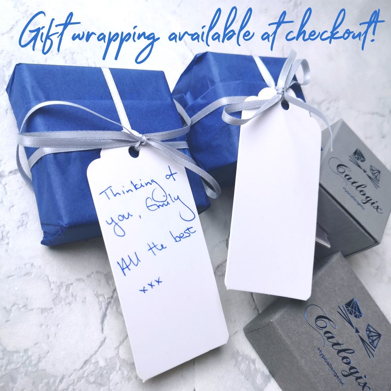 Op de afbeelding: Twee cadeaudozen verpakt in blauw papier met zilveren lint en een wit cadeaukaartje met een handgeschreven bericht. Het cadeaukaartje zegt "Thinking of you, Emily All the best xxx". De dozen staan op een wit oppervlak.
