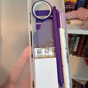 May include: A translucent plastic rectangular case containing stationery items including a roll of purple washi tape, a purple pen, a white pen with a light purple cap, a heart-shaped purple sticky note, and several silver metal paper clips.