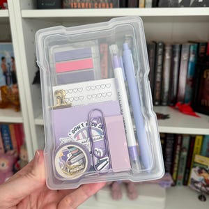 May include: A clear plastic container filled with stationery items. The container holds purple and pink sticky notes, pens, a paper clip, stickers, and a small notebook. The items are arranged neatly inside the container, creating a visually appealing set.