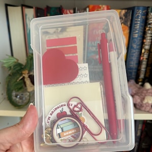 May include: A clear plastic rectangular storage case containing stationery items including a red pen, a white pen, a red heart-shaped sticky note, a stack of red adhesive tabs, a sheet of star stickers, a large red paperclip, and a roll of washi tape.